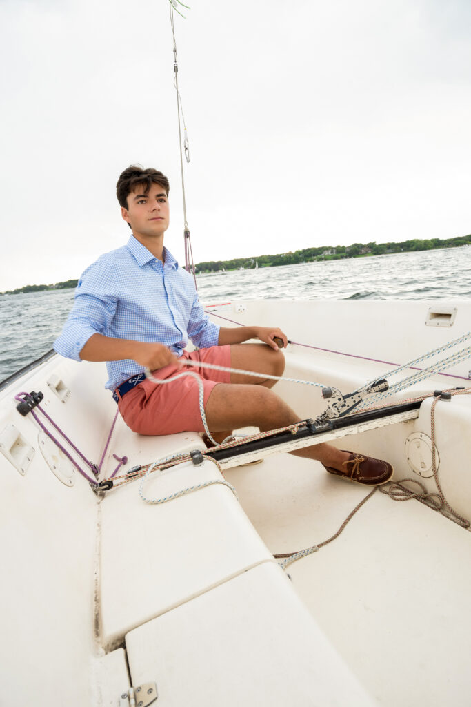 High school senior boy sitting on a sailboat in a blue button-down shirt during his senior portrait session.