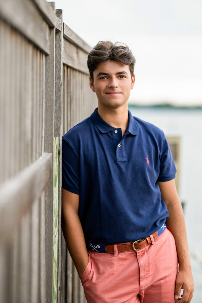 High school senior boy in a navy polo shirt posing beside a wooden fence near the water in Edina, Minnesota by Elissa Bean Photography.
